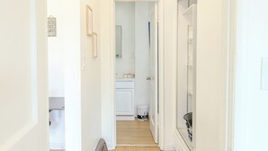Un couloir étroit aux murs blancs et au plancher en bois, qui mène à une salle de bain équipée de toilettes, d’un lavabo et d’un miroir.