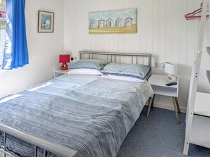 Ein ordentlich bezogenes Bett mit einer gestreiften Tagesdecke, ein Nachttisch mit einer Lampe, ein Bild mit Strandhütten und ein Fenster mit blauen Vorhängen.
