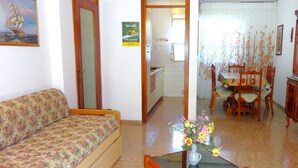 Una sala de estar con sofá, una mesa de comedor con sillas, una zona de cocina y una puerta que da a otra habitación.
