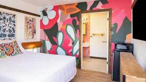 Une chambre à coucher avec un lit, une table de chevet en bois, un ventilateur de plafond et une fresque murale abstraite aux couleurs vives.