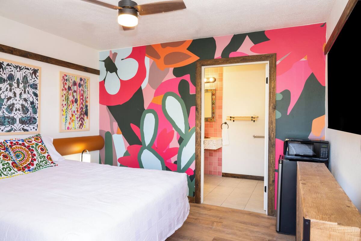 A bedroom with a bed, a wooden nightstand, a ceiling fan, and a colorful abstract wall mural.