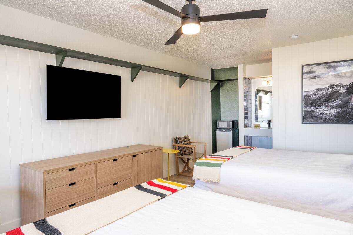 A bedroom with two beds, a TV, a ceiling fan, and a kitchen area visible through an open door.