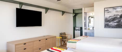 A bedroom with two beds, a TV, a ceiling fan, and a kitchen area visible through an open door.