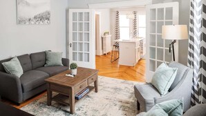 Una sala de estar con un sofá gris, una mesa de centro de madera y un sillón gris. Hay un cuadro en la pared y una lámpara sobre una mesita auxiliar.