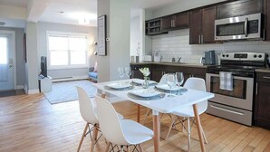 Cocina moderna con armarios de madera oscura, electrodomésticos de acero inoxidable y un comedor con sillas blancas y una mesa dispuesta para cuatro.