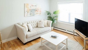 A living room with a white sofa, a coffee table, a flat-screen TV on a stand, a large window, and a potted plant. - 67 - Walking distance to downtown (Charlottetown)