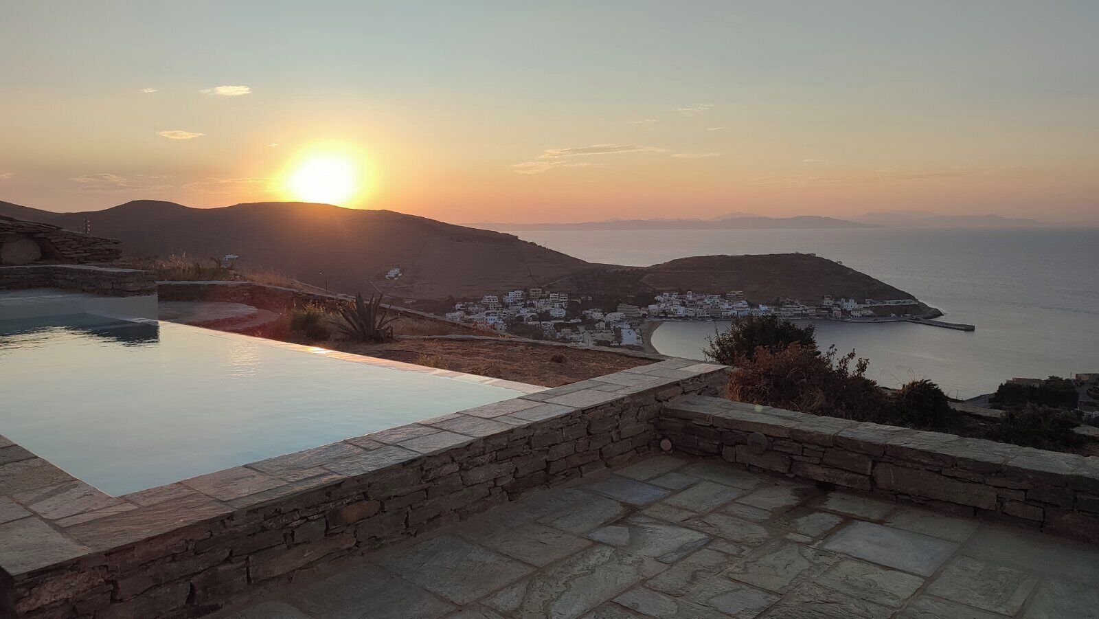 A stone pool with a view of a coastal town and mountains at sunset.
