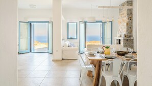 Ett rymligt vardagsrum med ett matbord för fyra personer, en vit soffa och utsikt över havet genom stora glasdörrar.