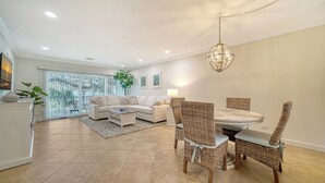 A modern living room with a sofa, dining table, and chairs. There is a large window with blinds, a hanging light fixture, and a plant.
