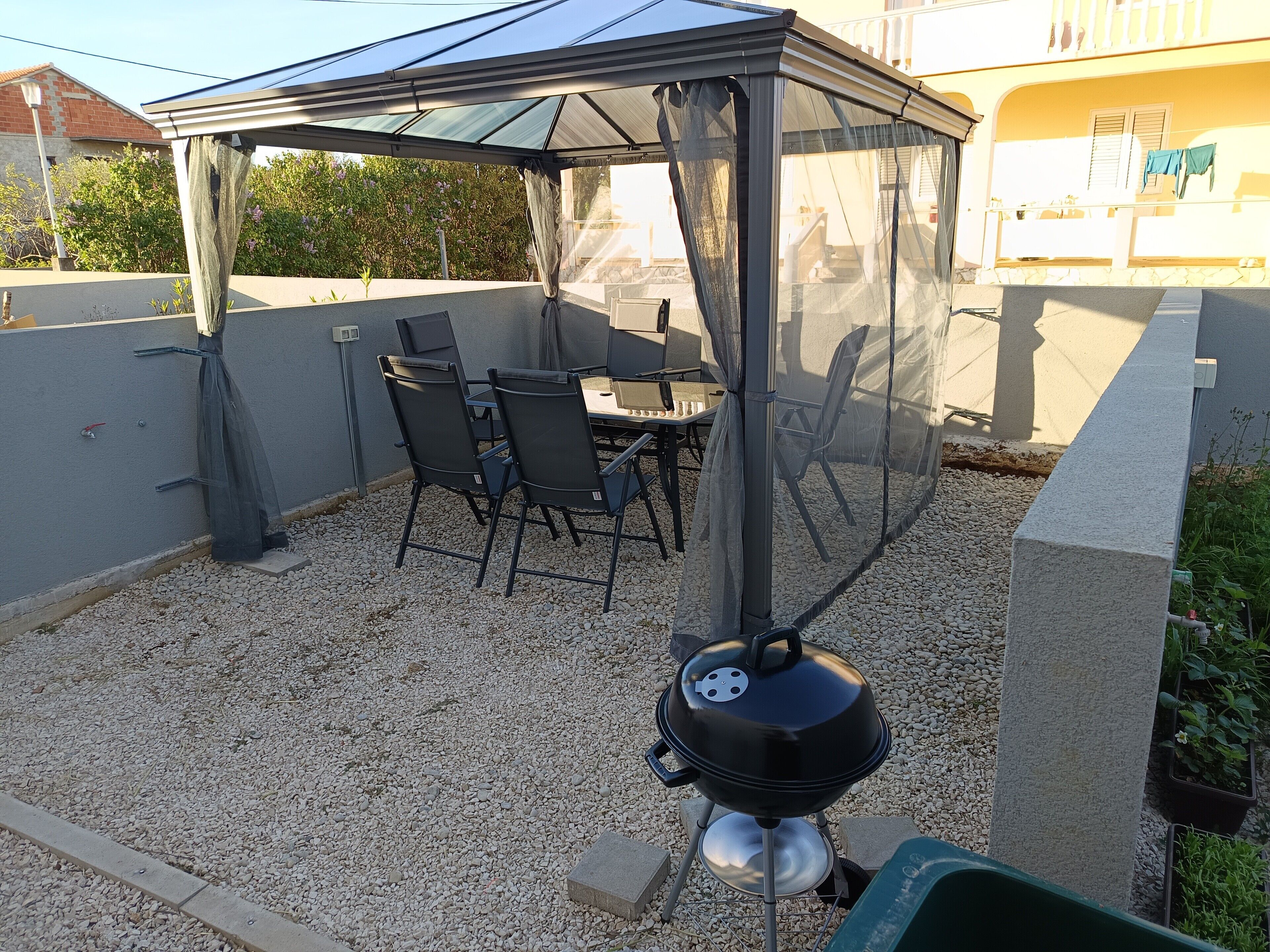Een overdekt terras met een barbecue, buitentafel en stoelen.