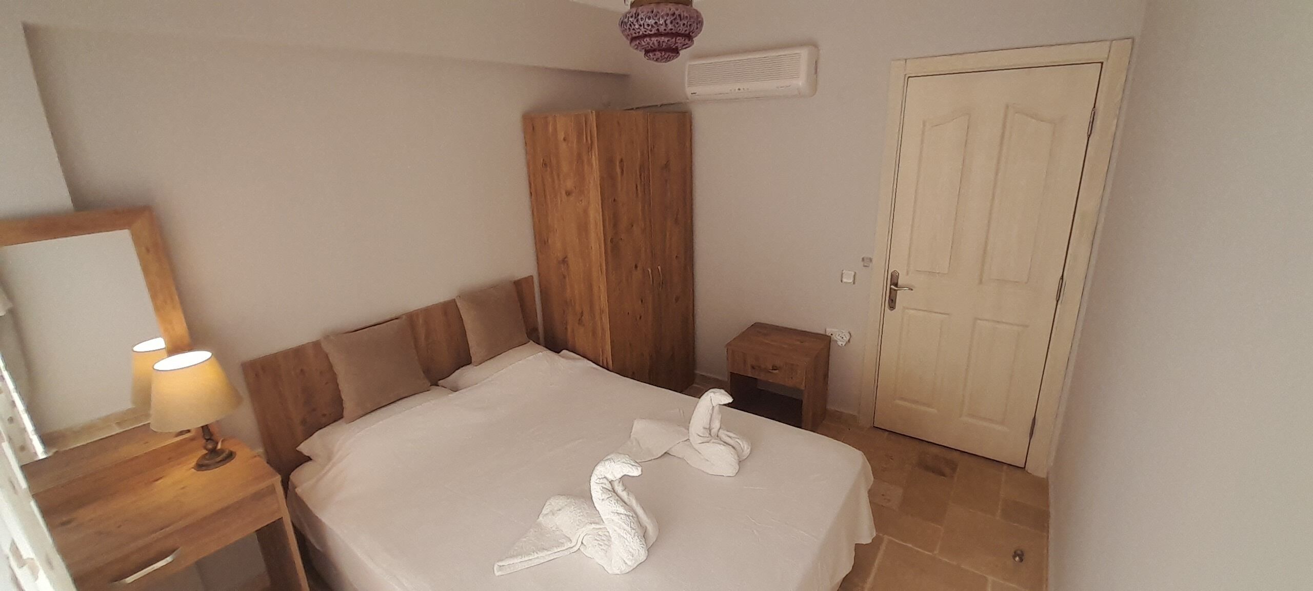 A bedroom with a bed, a wooden wardrobe, a bedside table with a lamp, a towel sculpture, and a door.