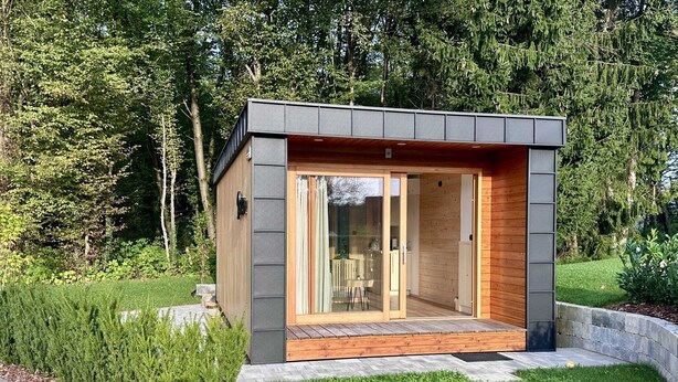 Una cabaña moderna y compacta con puerta de cristal, rodeada por un jardín cuidado y un sendero de piedra que lleva a la entrada.