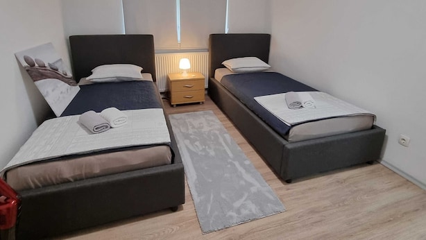 A compact room with two single beds, a small bedside table with a lamp, a wooden floor, and a window with blinds. - Charming Rustic Apartments Brest - One Bedroom Apartment (Ig)