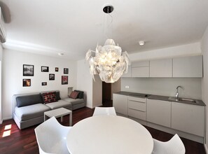 Una sala de estar moderna con un sofá gris, una mesa de comedor blanca y una zona de cocina con gabinetes grises y un fregadero de acero inoxidable.