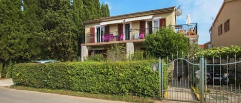 A two-story residential building with a balcony, a gated entrance, and a well-maintained hedge.