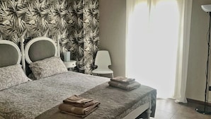 En snyggt bäddad säng med ett mönstrat överkast, två sängbord med lampor, en stol och ett stort fönster med tunna gardiner.