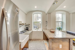 Uma cozinha moderna com uma geladeira grande, bancadas de madeira e um padrão de piso em herringbone. Há duas janelas com arcos e uma ilha de madeira com uma tigela sobre ela.