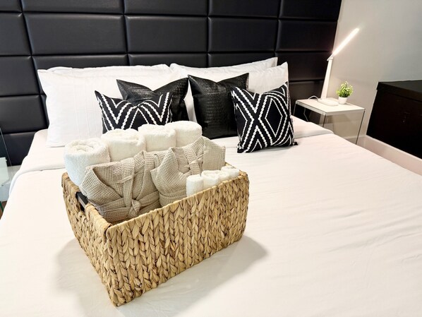 A neatly made bed with a woven basket filled with towels and pillows, a bedside table with a lamp, and a dark headboard.