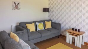 A modern living room with a grey sofa, yellow cushions, a small wooden table, and a wall-mounted TV. - Rosedene Cottage - UK48604 (Chapel St. Leonards)
