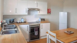 A compact kitchen with white cabinets, a stainless steel oven, a microwave, and a toaster. There's a wooden table with a chair, and a white refrigerator. - Rosedene Cottage - UK48604 (Chapel St. Leonards)