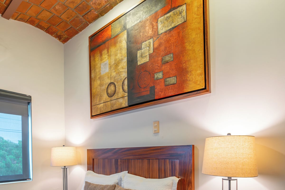 Cama bem arrumada com cabeceira de madeira, duas mesas de cabeceira com luminárias, e um grande quadro abstrato acima da cama.