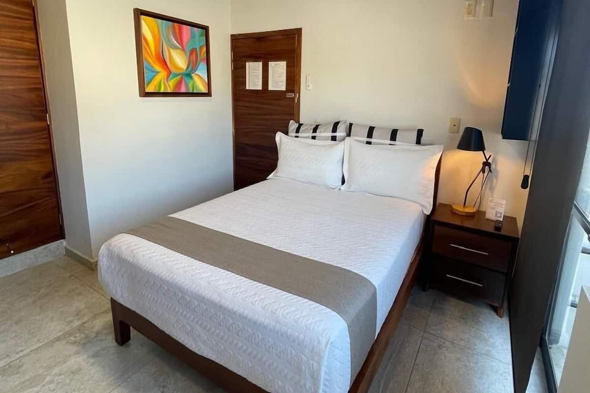 Quarto com cama, mesa de cabeceira, um quadro, porta de madeira e luminária no teto.