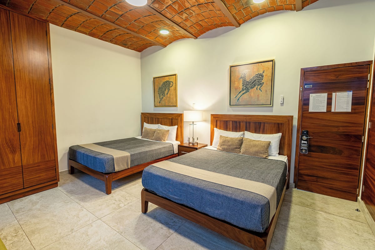 Quarto de hotel com duas camas, cabeceiras de madeira, teto de tijolos e guarda-roupa de madeira.