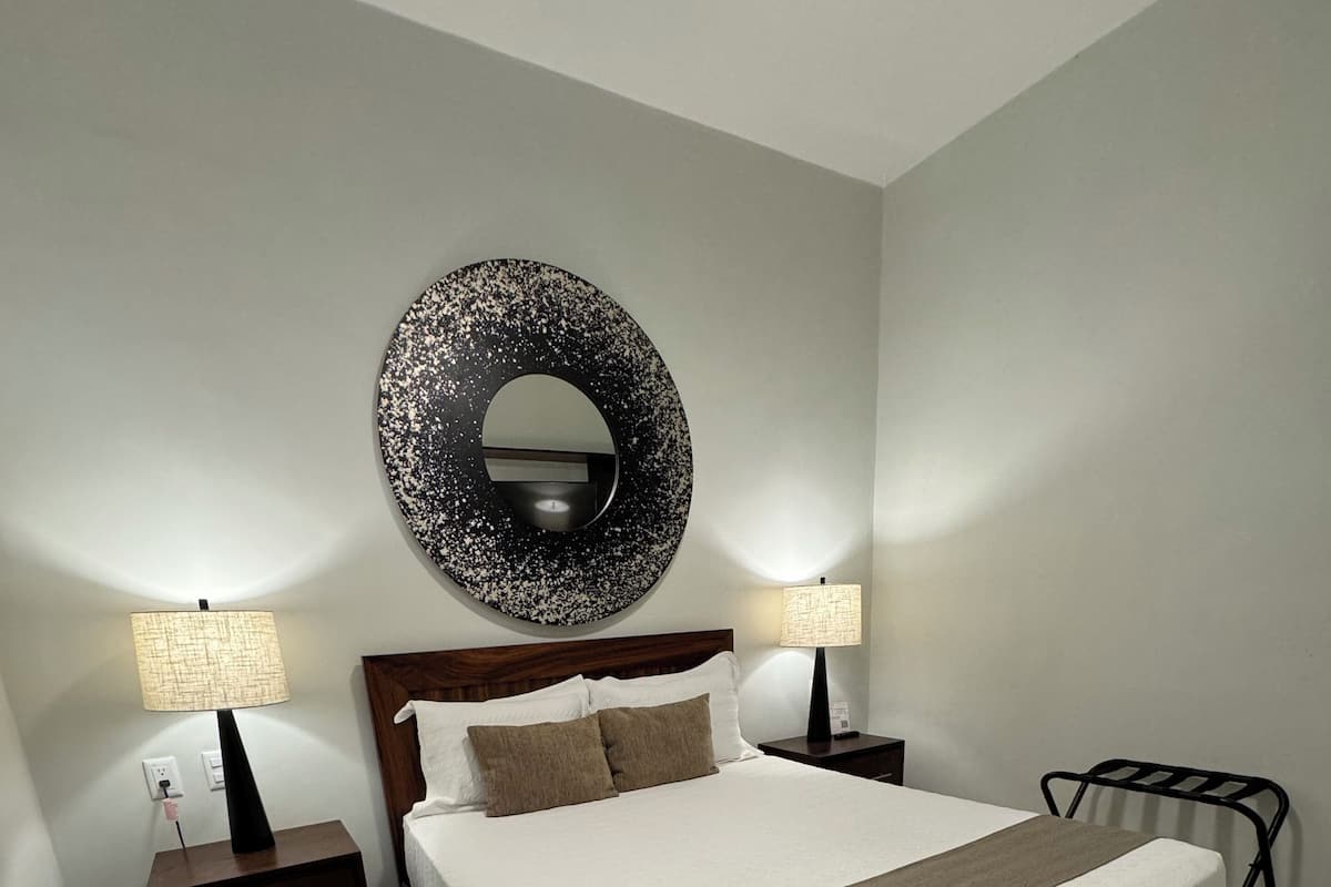 Uma cama bem arrumada com cabeceira de madeira, duas mesas de cabeceira com abajures, um espelho redondo na parede e uma cadeira de metal.