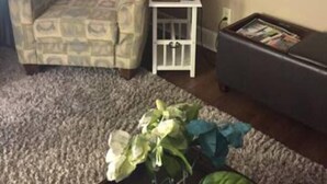 A living room with a patterned armchair, a small white side table with a lamp, a dark sofa, and a carpeted floor.