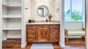 A bathroom with wooden cabinets, a round mirror, and a window with a view of greenery. - Modern Farmhouse with Pool & Vineyard (Santa Rosa)