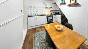 A compact kitchen with white cabinets, a black countertop, and a wooden dining table with a bowl of fruit. - Beach Oasis Cottage - UK48530 (Margate)