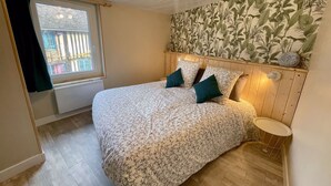 Ein Schlafzimmer mit einem blumigen Bettüberwurf, einem Holzkopfteil, einem Fenster mit Jalousien, einem kleinen runden Tisch und einer Wand mit tropischer Pflanzen-Tapete.