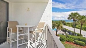A balcony with a white table and chairs, a small window, and a view of a road with palm trees and a clear sky.