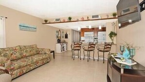 A living room with a sofa, a dining area with chairs, a kitchen in the background, and a TV mounted on the wall.