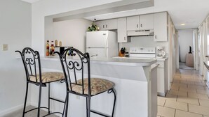 A modern kitchen with white cabinets, a central island, and two bar stools. - Cape Winds #211 Ocean view 1 bedroom condo (Cape Canaveral)