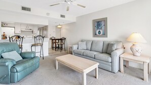 A living room with a sofa, a coffee table, a lamp, and a ceiling fan. - Cape Winds #211 Ocean view 1 bedroom condo (Cape Canaveral)