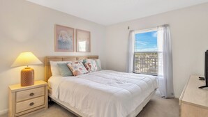 A bedroom with a bed, nightstand, lamp, and a window with curtains. - Cape Winds #211 Ocean view 1 bedroom condo (Cape Canaveral)