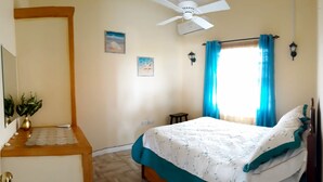 Une chambre à coucher comprenant un lit, un ventilateur de plafond, une fenêtre avec des rideaux, une petite table et une lampe fixée au mur.
