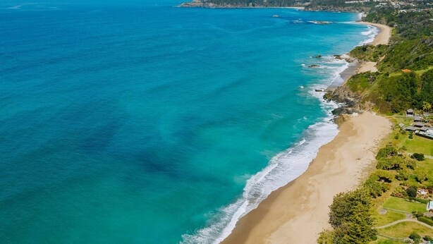 Vue aérienne d’une zone côtière avec une plage de sable, des eaux turquoise et limpides, et une colline densément végétalisée où se trouvent des bâtiments.