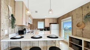 A modern kitchen with wooden walls, a central island with stools, hanging pendant lights, and a view of a house through a window.