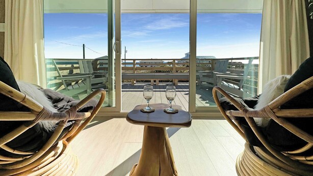 A balcony with wicker chairs and a small table, overlooking a coastal area with buildings and a clear sky.
