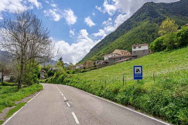 Una carretera rural con casas en una ladera, un cartel que indica un núcleo de turismo rural, y un cielo despejado.