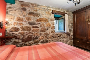 A room with a stone wall, a bed with a plaid bedspread, a wooden wardrobe, and a window with a curtain.