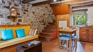 A rustic interior with a stone wall, wooden staircase, a sofa with blue cushions, a dining table with chairs, and a kitchen area.