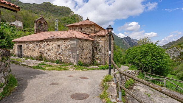 A stone building with a tiled roof, a bell tower, and a paved road leading to it.