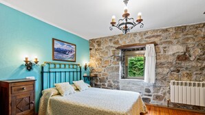 A bedroom with a stone wall, a bed with a patterned bedspread, a wooden dresser, and a window with a view of greenery.