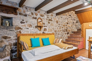 Une chambre aux murs de pierre, avec un lit en bois, une étagère en pierre et un escalier en bois.
