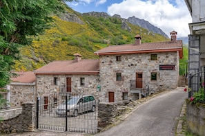 Un edificio de piedra con techo de tejas rojas, un coche estacionado al frente y montañas al fondo.