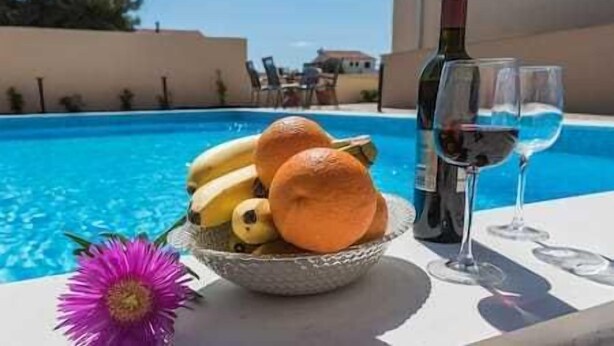 A poolside table with a bowl of fruit, two wine glasses, and a bottle of wine.