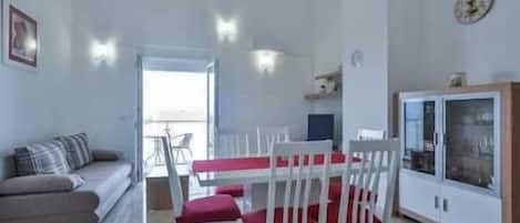 A spacious living area with a sofa, dining table, and chairs. There is a balcony visible through an open door.
